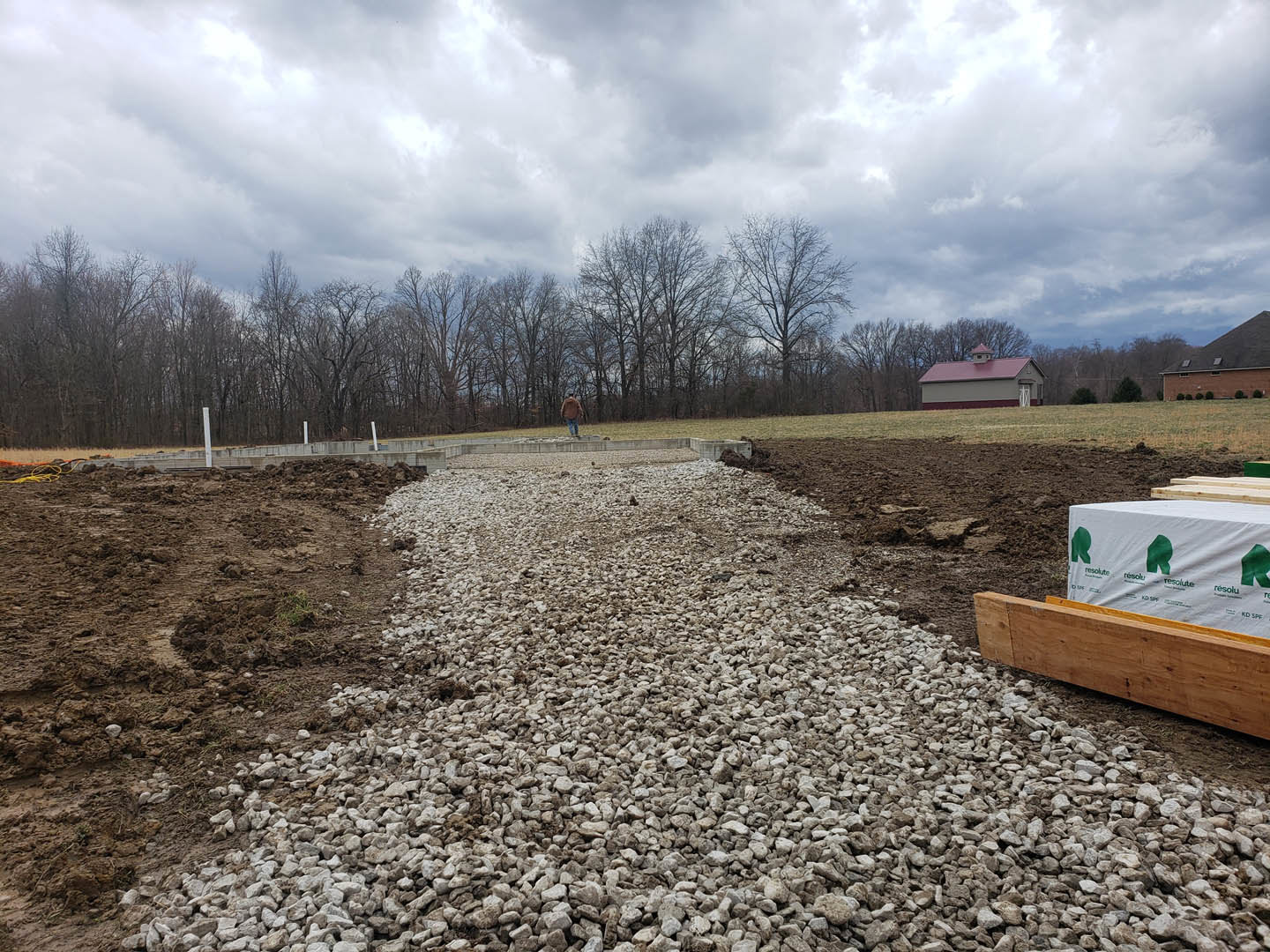 Gravel driveway bordered by trees under a cloudy sky, wooden board on ground with white container placed on top, small chimney visible on roof in background