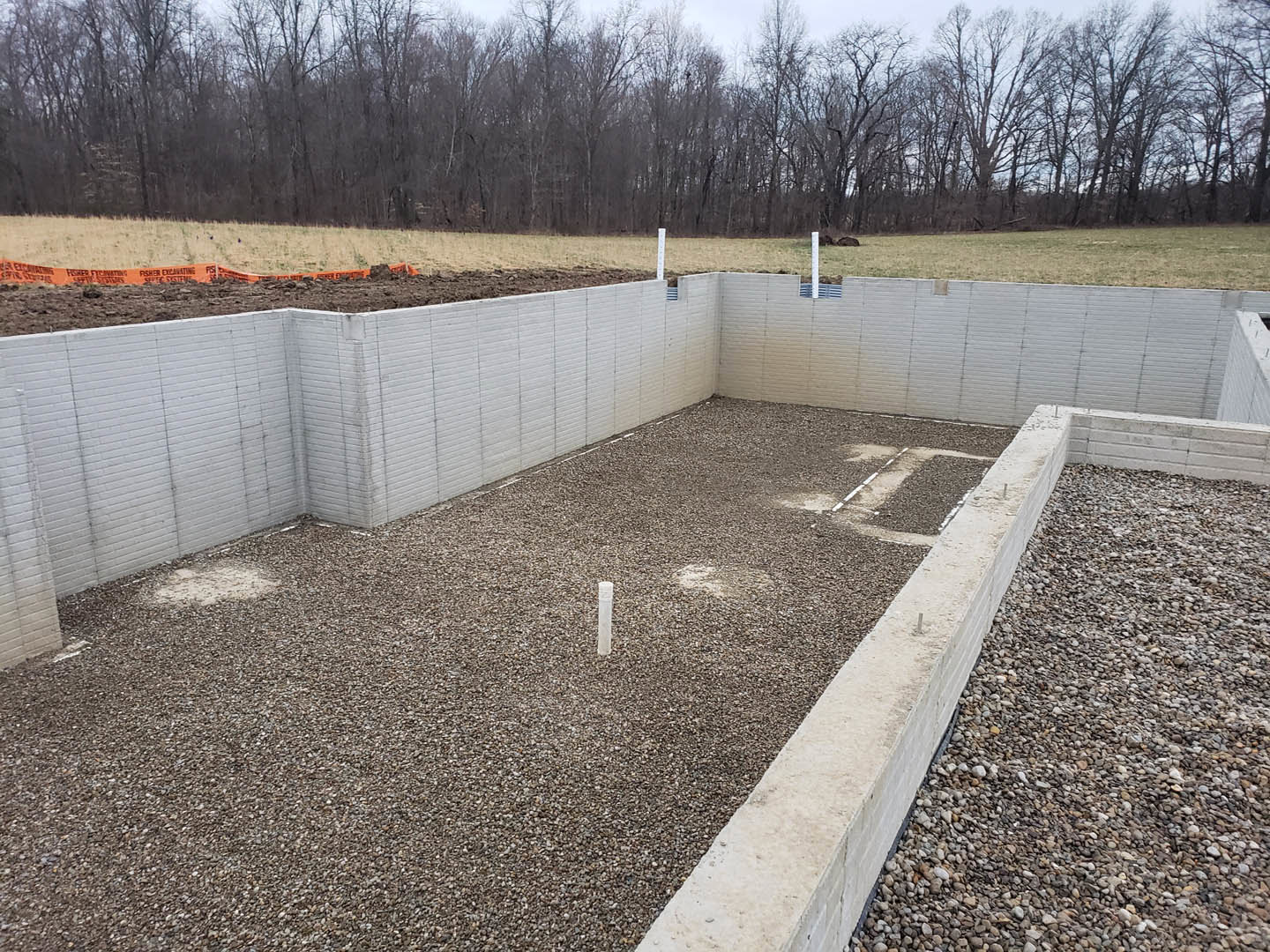 Concrete foundation bordered by gravel, enclosed by a wooden fence, with trees visible in the background and a metal pole set in the gravel area