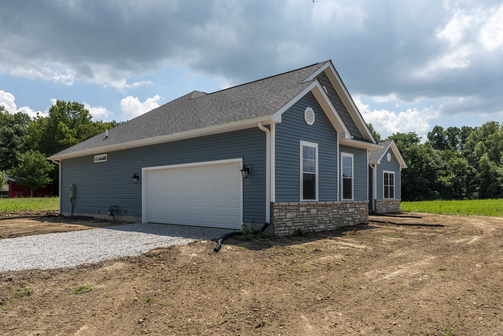 White garage with gray roof and stone wall, black pipe in dirt beside garage, window with white frame, American Gothic House visible in background, surrounded by trees and cloudy