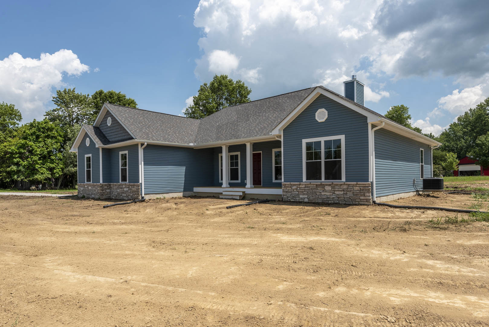 Modern house with white-framed windows, covered porch, black metal-framed object near dirt yard, exposed pipe, and mature tree under cloudy sky