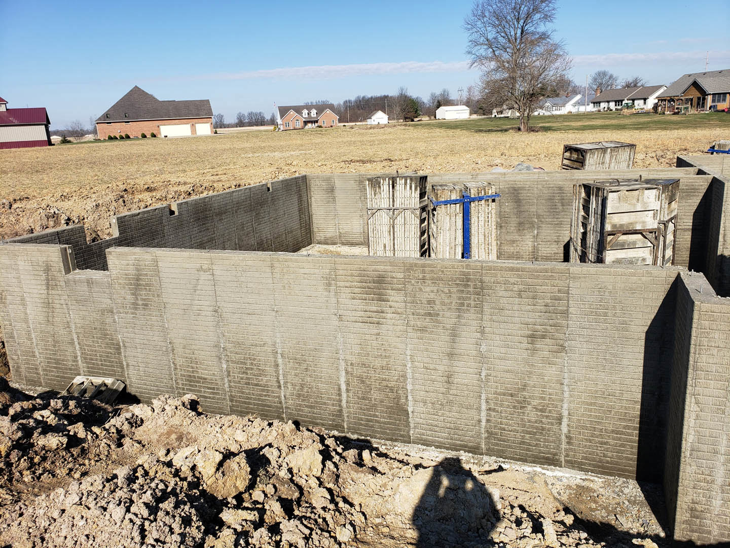 Concrete foundation slab with exposed soil, surrounded by grassy field, large tree, brick house with garage door, red-roofed barn, and blue sky with scattered clouds