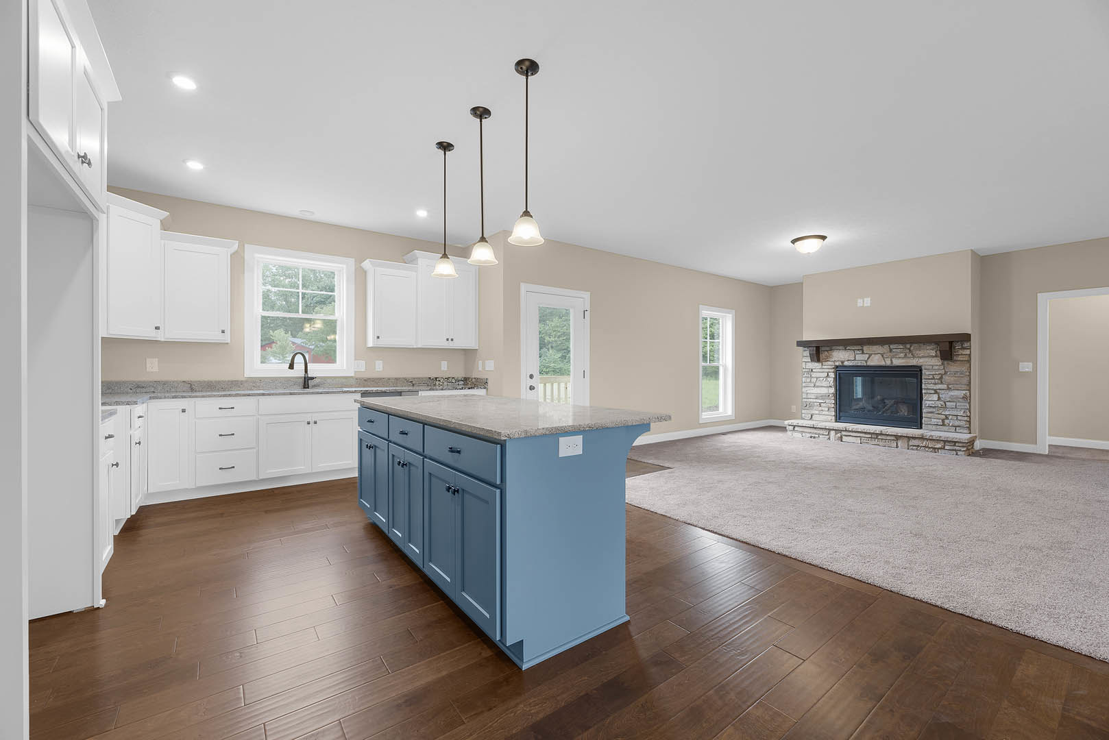 Open-concept kitchen and living room featuring a marble-topped island, white cabinetry, glass-door fireplace, stainless steel sink beneath a window overlooking trees, and neutral