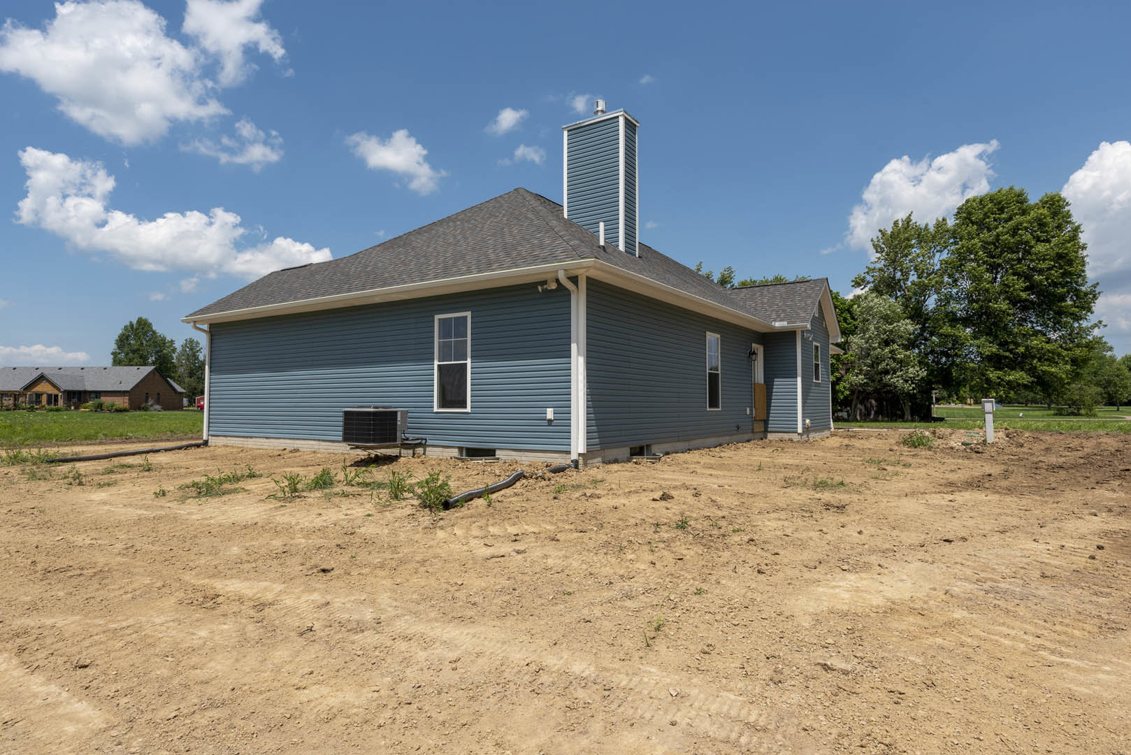 Two-story house with grey roof, white-framed windows, and brick chimney, set behind a dirt field with exposed pipe; black grill visible in foreground, surrounded by open land and