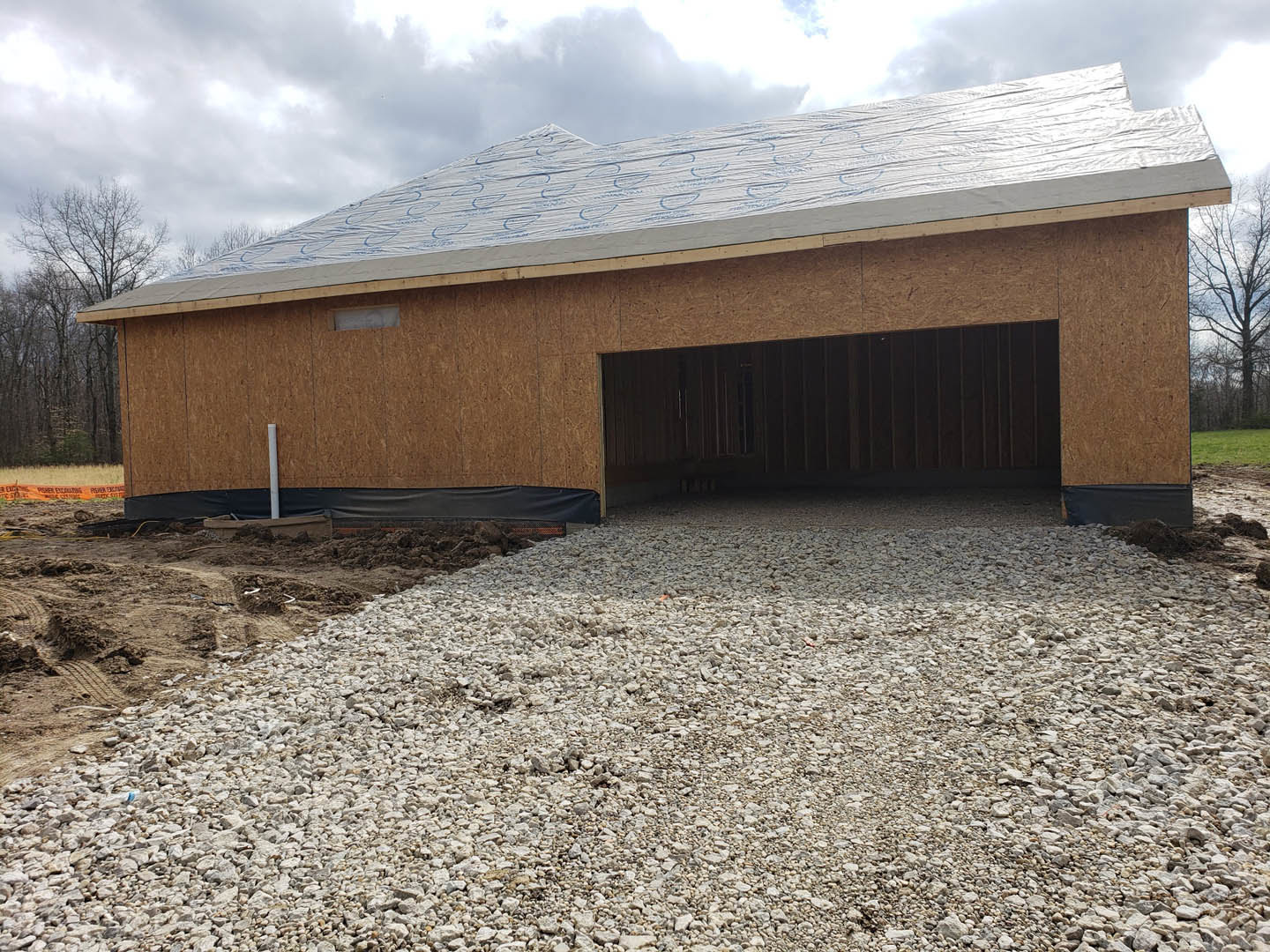 Modern home exterior with attached garage, gray roof, unfinished gravel driveway, and surrounding trees.
