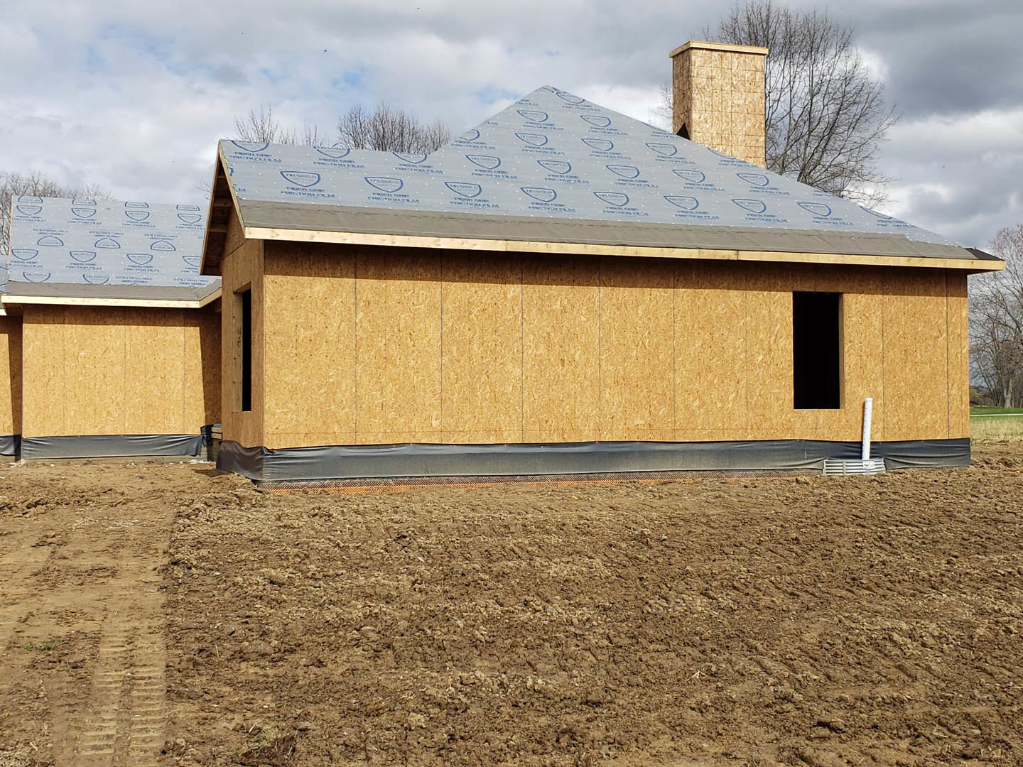 Wood-framed house under construction with exposed roof trusses, surrounded by dirt, trees, and temporary fencing