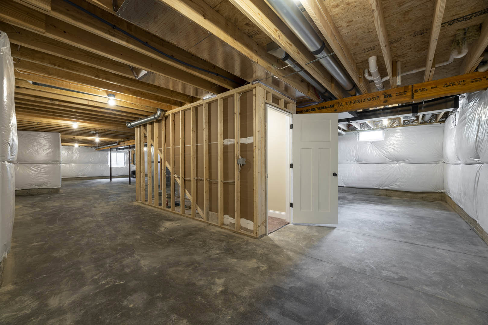 Concrete floor with exposed wood framing, open white door, white plastic wall covering, wooden beam marked with black text, plastered wall, and visible ceiling insulation.