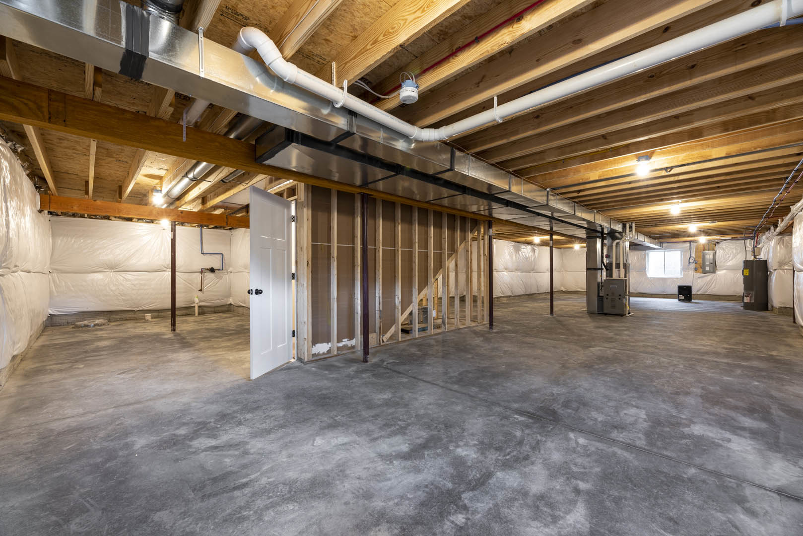 Concrete-floored basement with exposed ceiling pipes, white walls, and a closed door