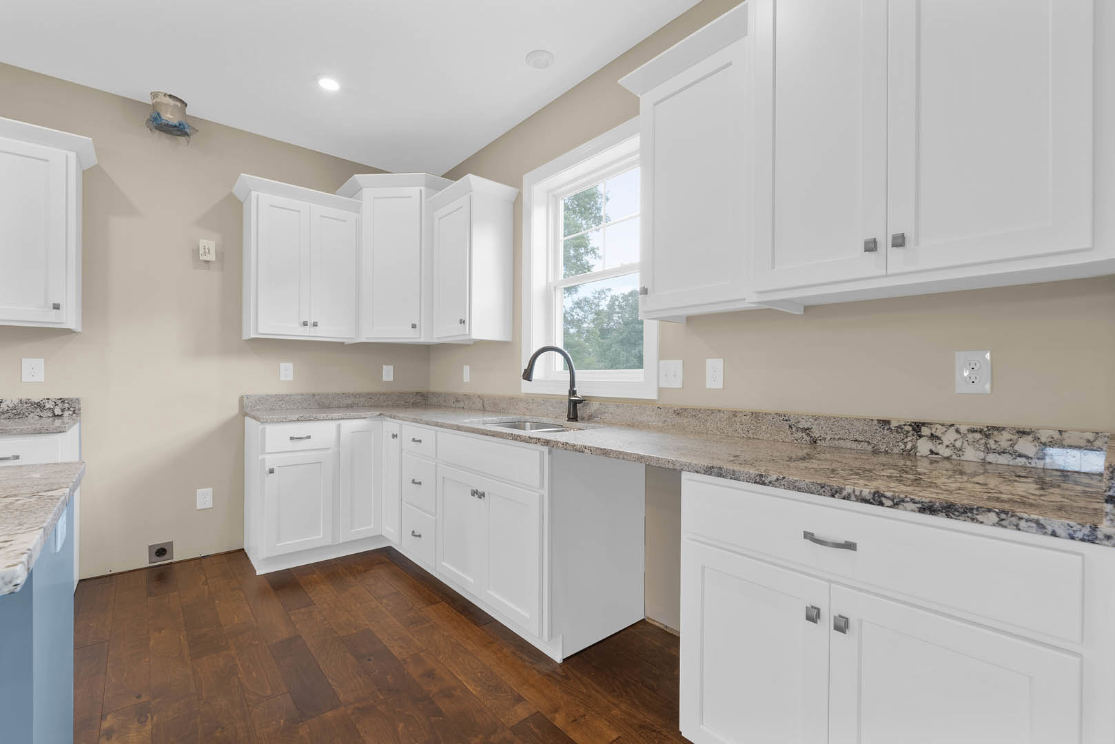 White kitchen with granite countertops, stainless steel faucet, white cabinets, wall outlet, and a white bucket with blue cloth on the floor.