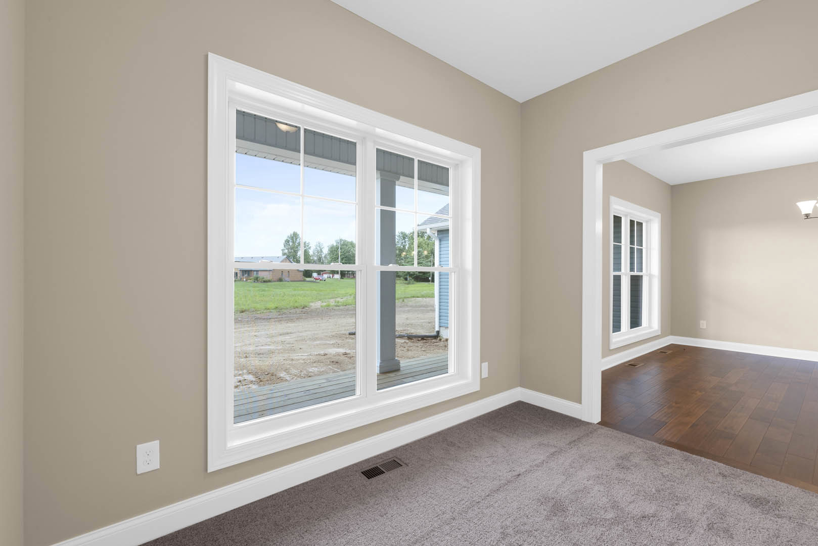 Carpeted bedroom with white walls, large window overlooking green yard, white electrical outlet visible on wall, simple baseboard molding