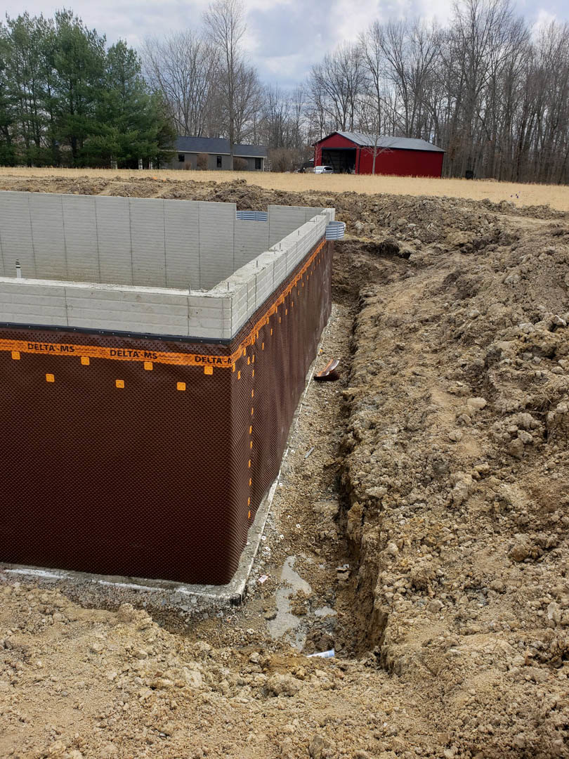 Concrete house foundation surrounded by dirt, with a red barn and leafy tree in the background under a partly cloudy sky