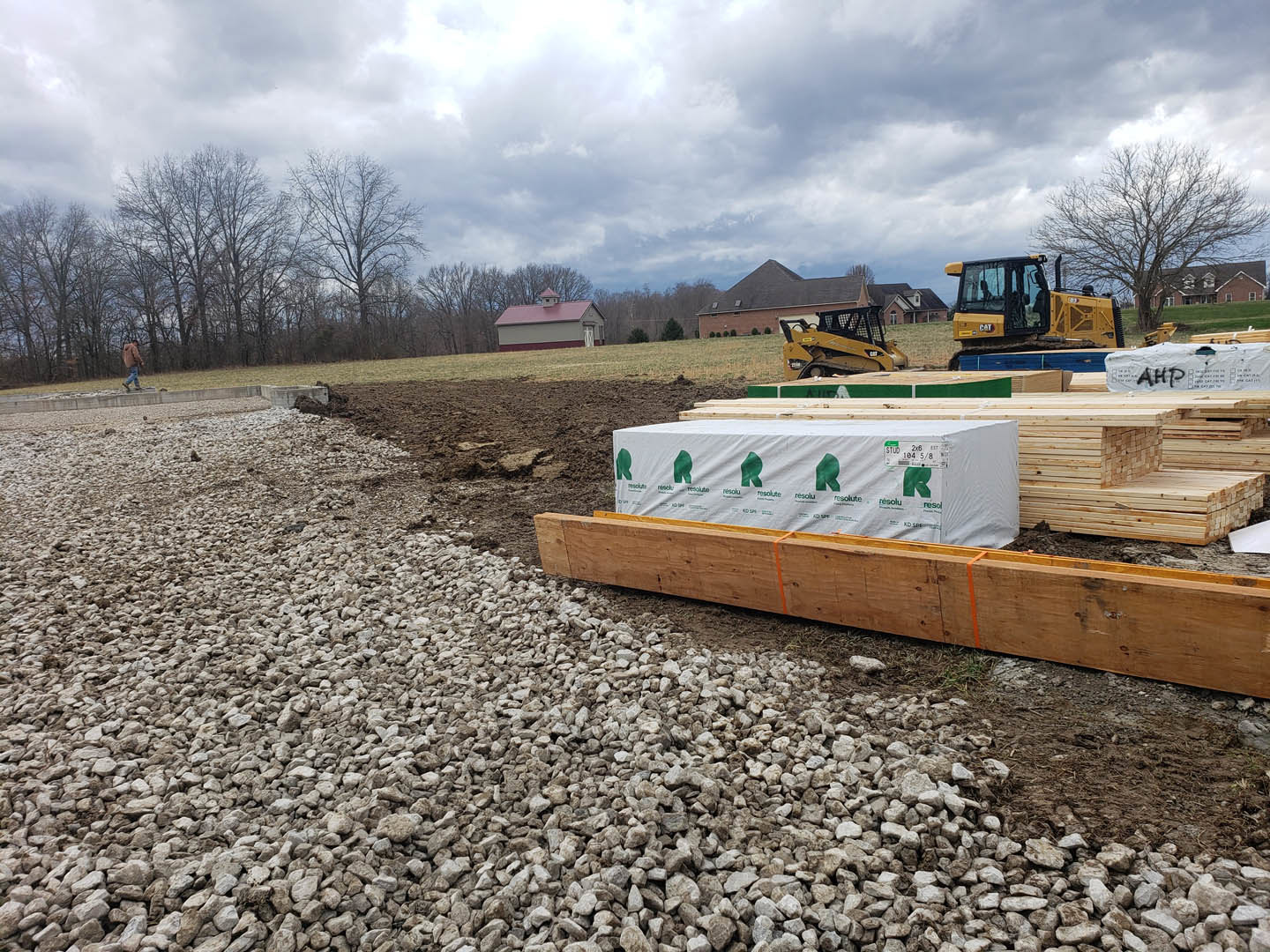 Construction site with stacked lumber, wooden box secured by orange straps, white tarp printed with green logos, yellow bulldozer, roof featuring a small chimney, and scattered