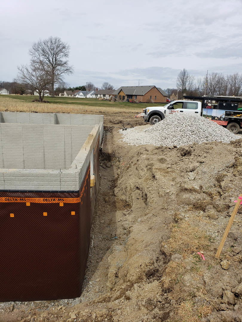 White pickup truck with red lettering parked beside a mound of gravel near a partially constructed building, surrounded by trees and open ground.