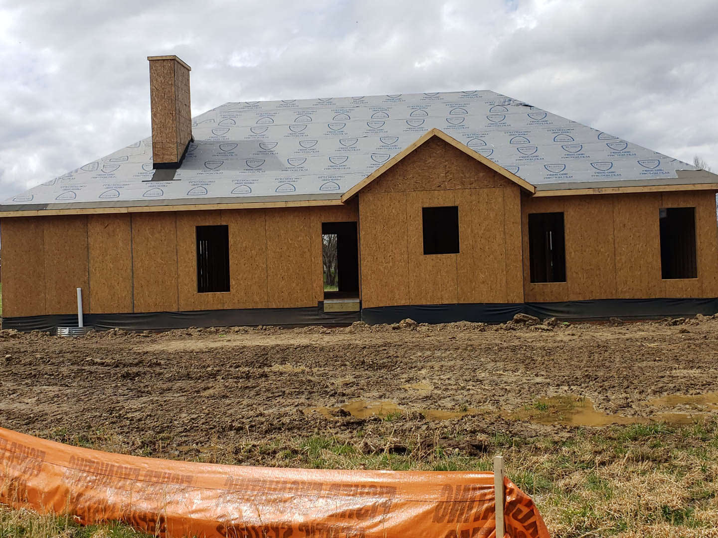Partially built house with grey roof, surrounded by wooden fence, dirt yard, and grassy area under cloudy sky