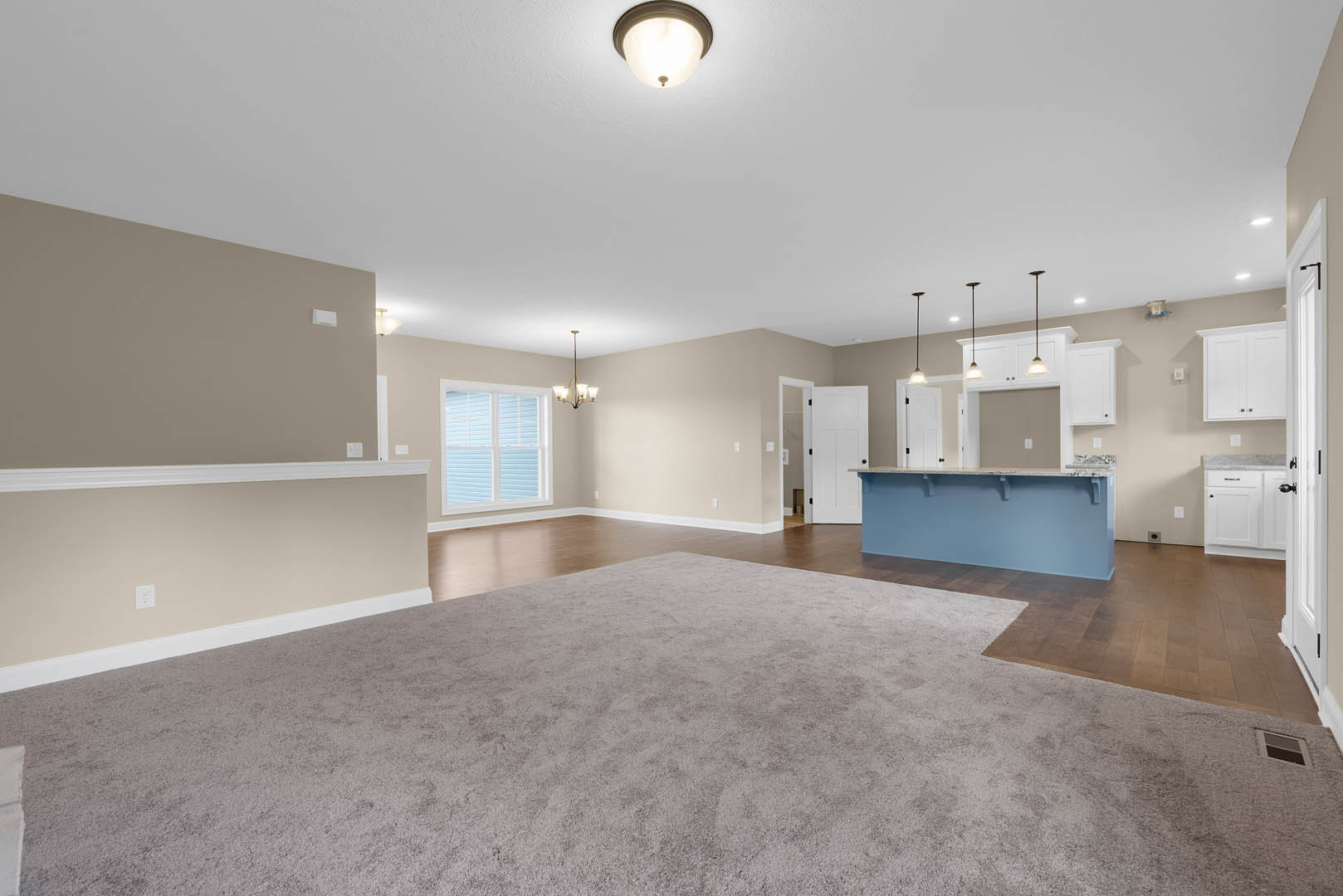 Open-concept living room with carpet flooring, blue accent wall featuring white shelves, adjacent kitchen and bar area, large window, modern ceiling light fixture, white door with