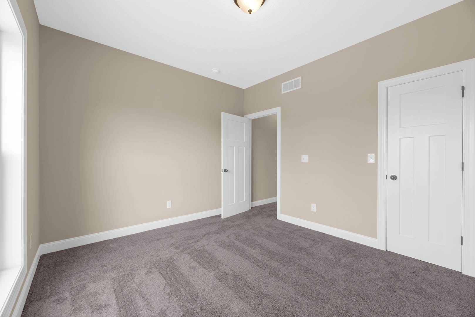 Beige-walled room with white door, silver knob, carpet flooring, white-framed window, and ceiling light fixture