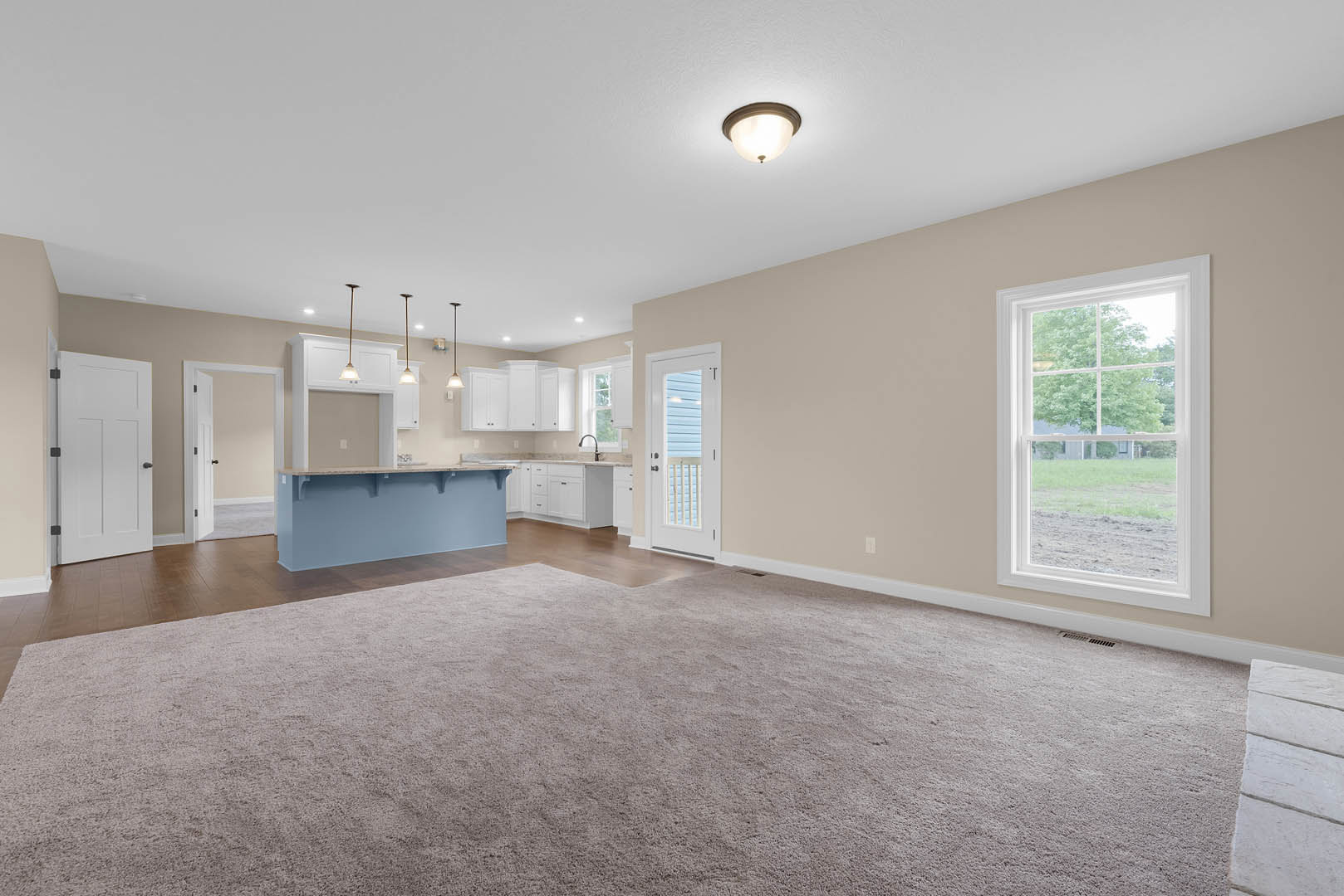 Open concept living room with carpet flooring, adjacent kitchen featuring tile floors and cabinetry, bar area, large window overlooking field and trees, white doors with black