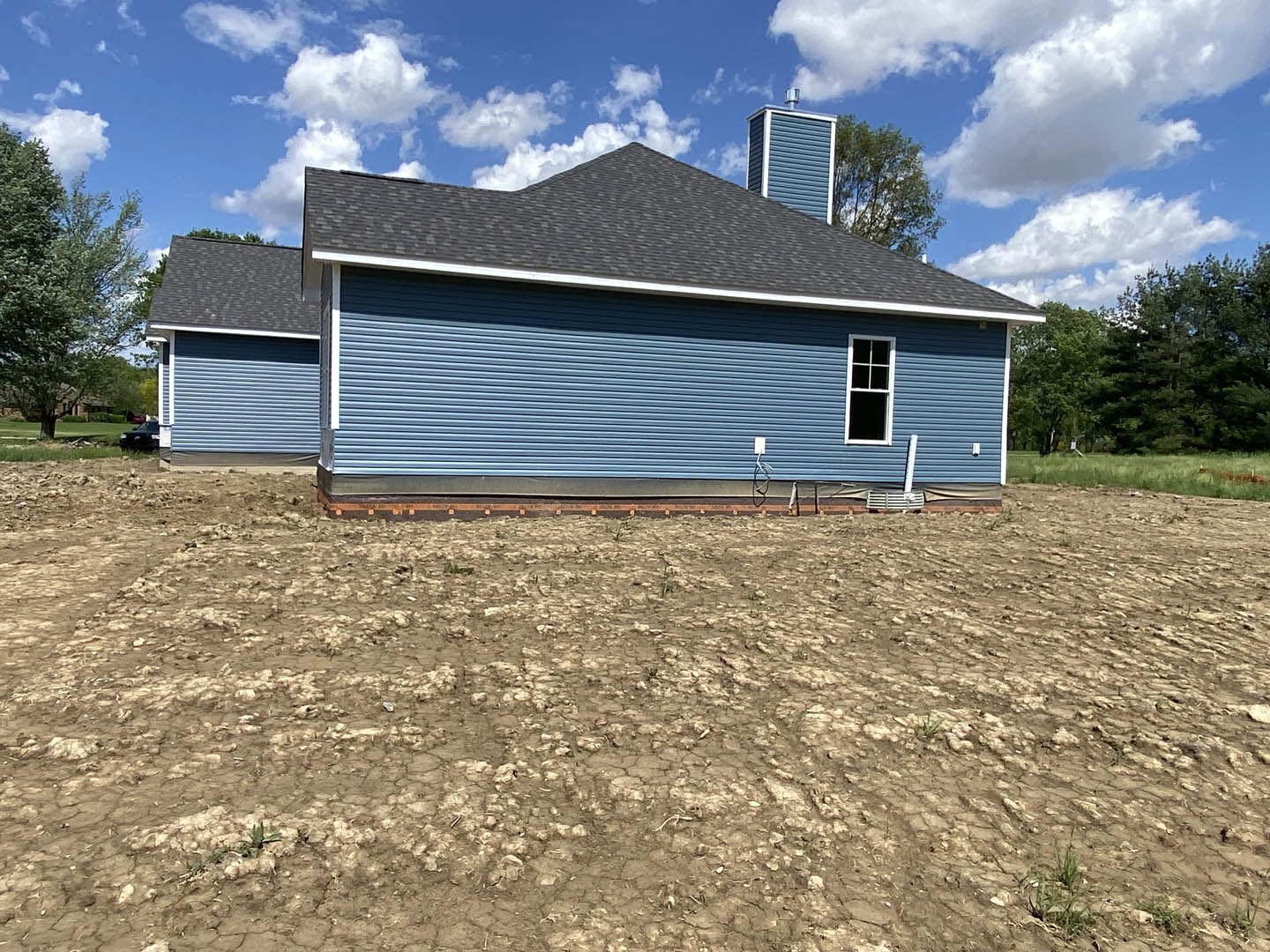 Blue house with white-framed windows and chimney, surrounded by a large field of dry, cracked dirt; leafy tree visible beside the home under a partly cloudy sky.