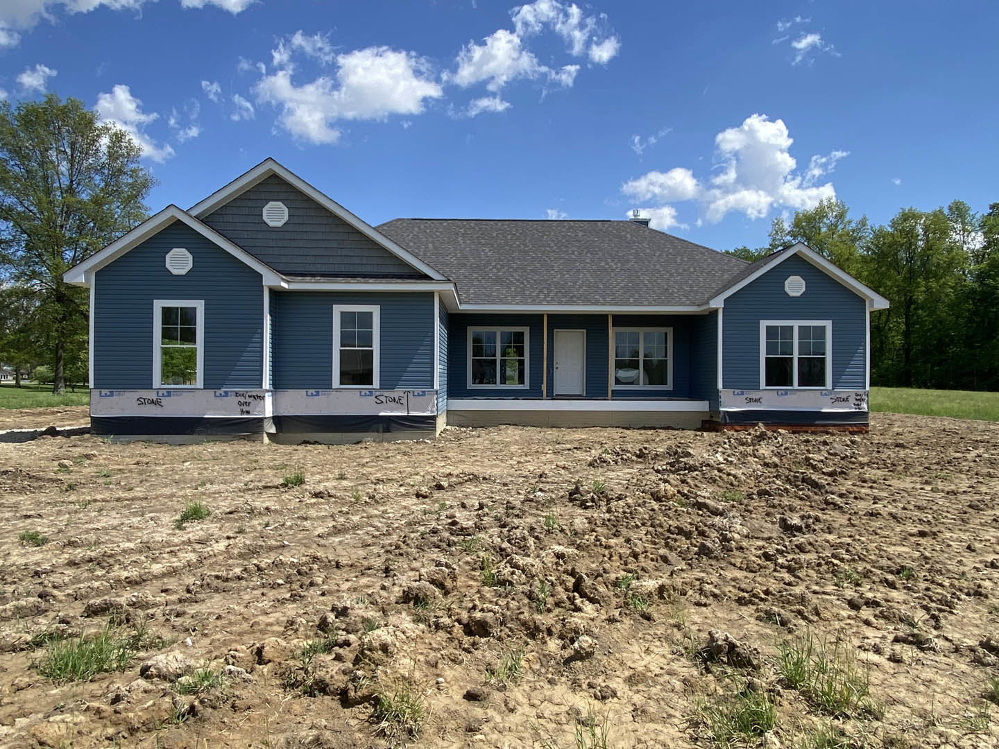 Blue-roofed house with white siding, white-framed windows, and a white front door, set behind a dirt yard with sparse grass; mature tree and cloudy sky in background.