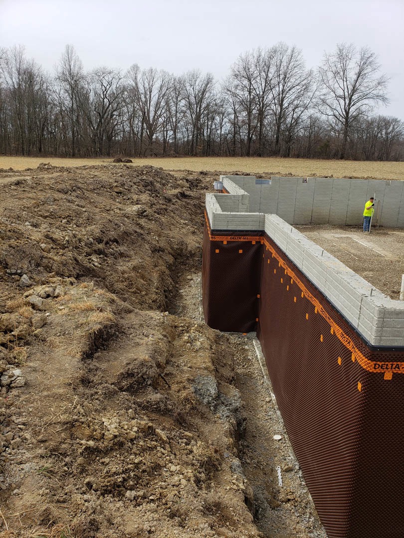 Concrete foundation wall with circular opening, surrounded by dirt trench and grassy ground, man standing in front, trees and sky in background