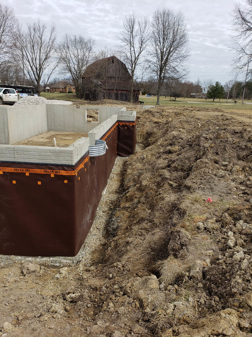 Concrete house foundation with exposed rebar, grey plastic pipe on brown soil, white truck with red writing parked nearby, barn and trees in background under partly cloudy sky