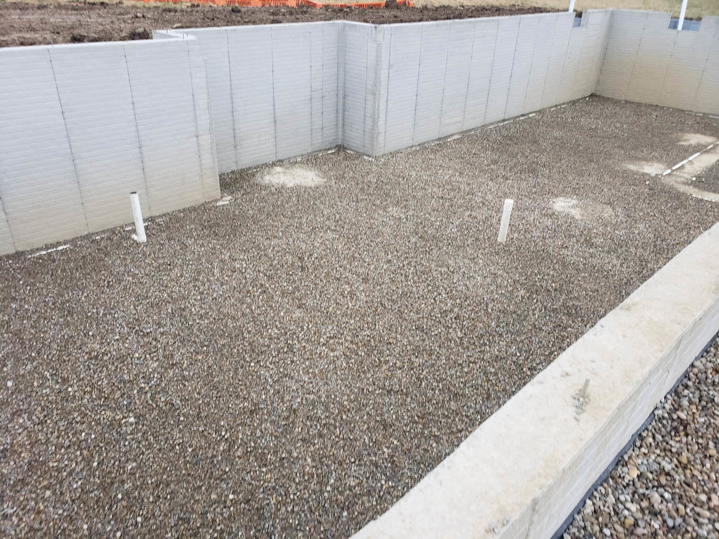 Concrete foundation with gravel floor, adjacent white walls, and a building in the background; dirt and gravel surround a sidewalk, with a concrete wall visible.