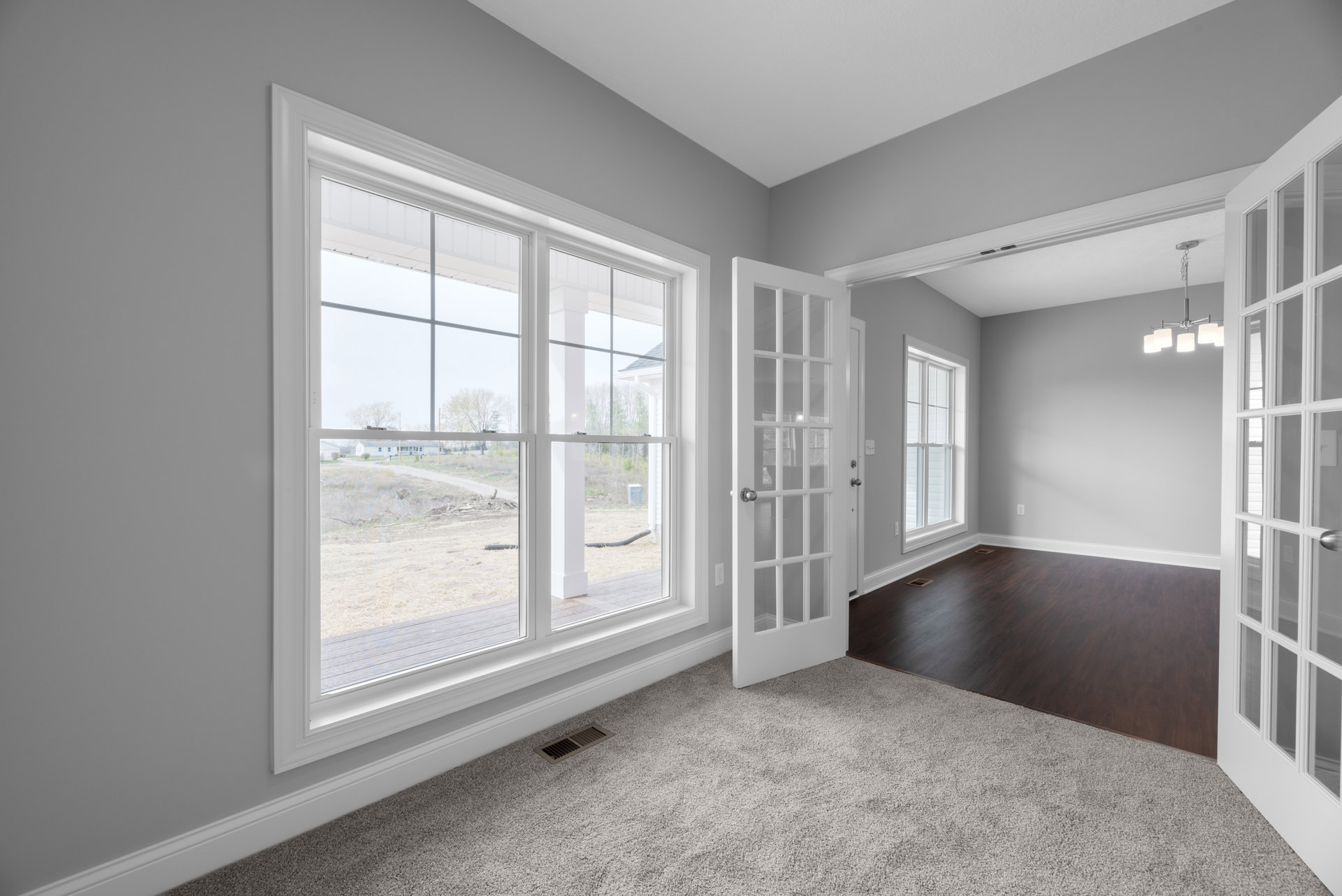 Carpeted room with white plaster walls, large windows with blinds, and a paneled door; windows overlook a yard, dirt road, and neighboring house.