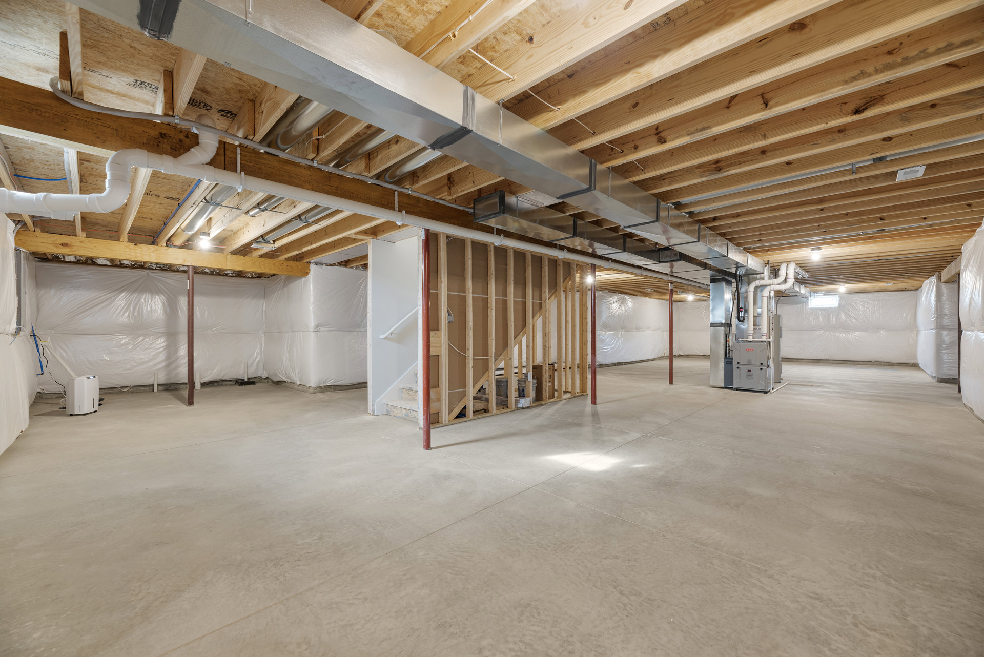 Basement room featuring exposed wood ceiling beams, visible metal pipes, white device with blue light, stairs along one wall, and a metal box with glass lid on the floor.