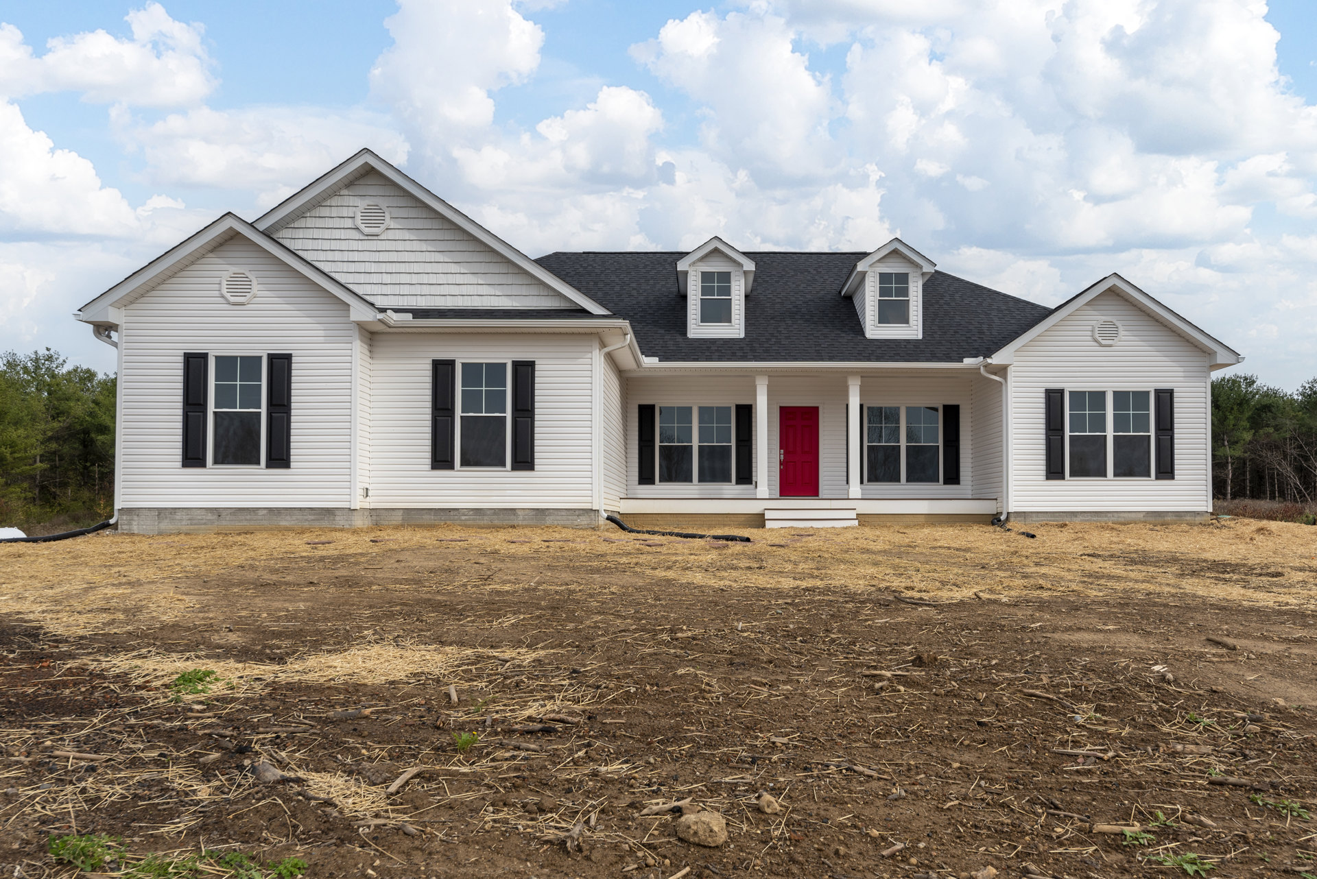 White siding house with black shuttered windows, red front door framed in white, gabled roof with dormer window, dirt yard with scattered sticks, cloudy sky overhead