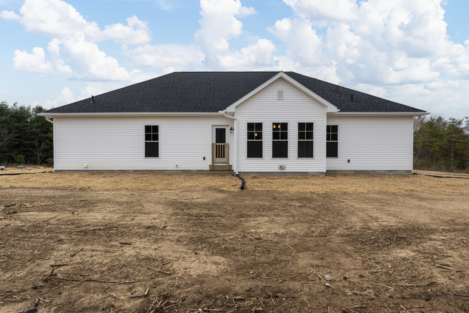 White house with black roof, wooden front door, black window panels, wooden gate set in white wall, dirt field with exposed pipe, blue sky and scattered clouds overhead