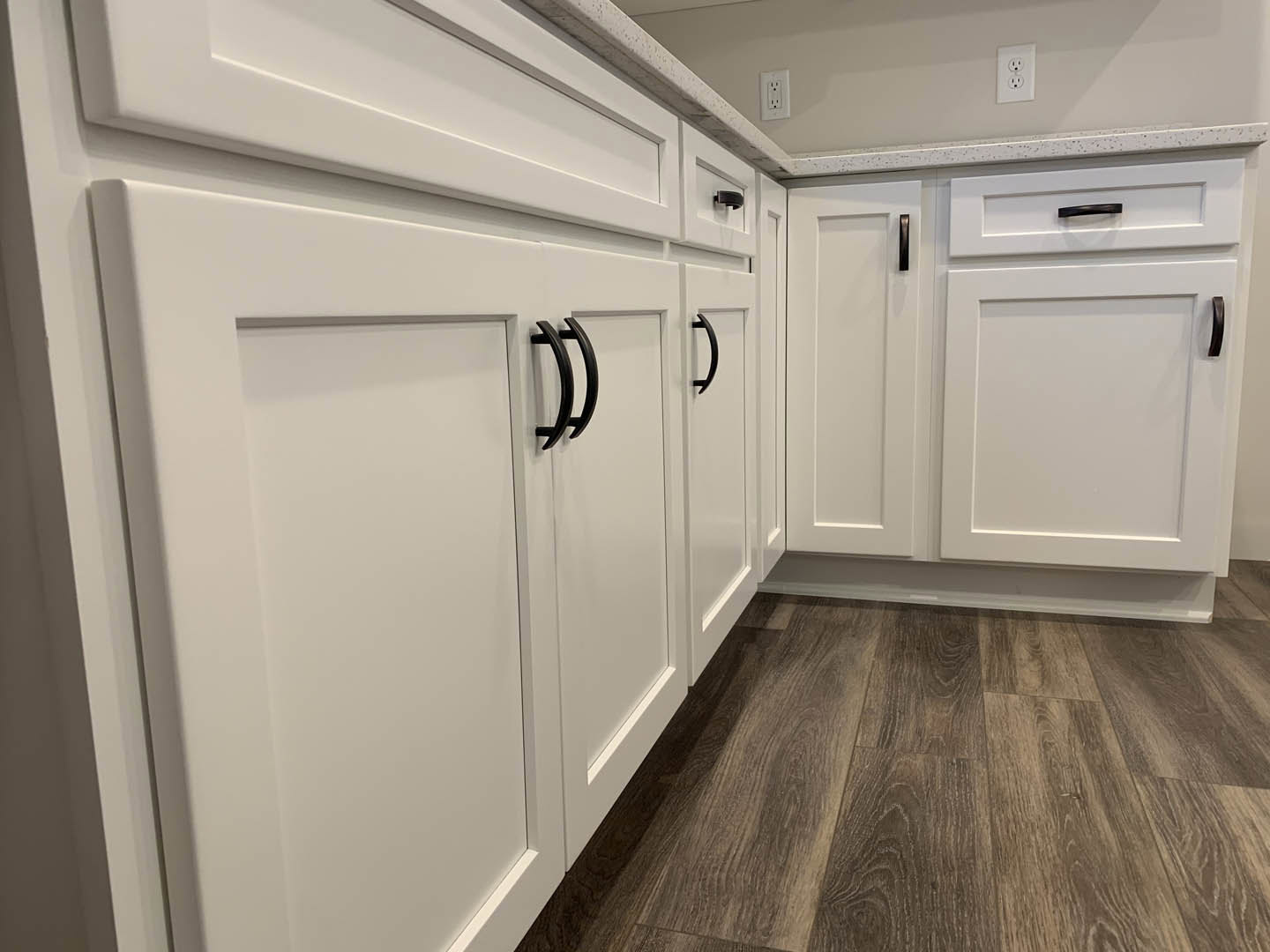 White kitchen cabinets with black handles, wood flooring, white electrical outlet with black dots, close-up of cabinet doors and wall.
