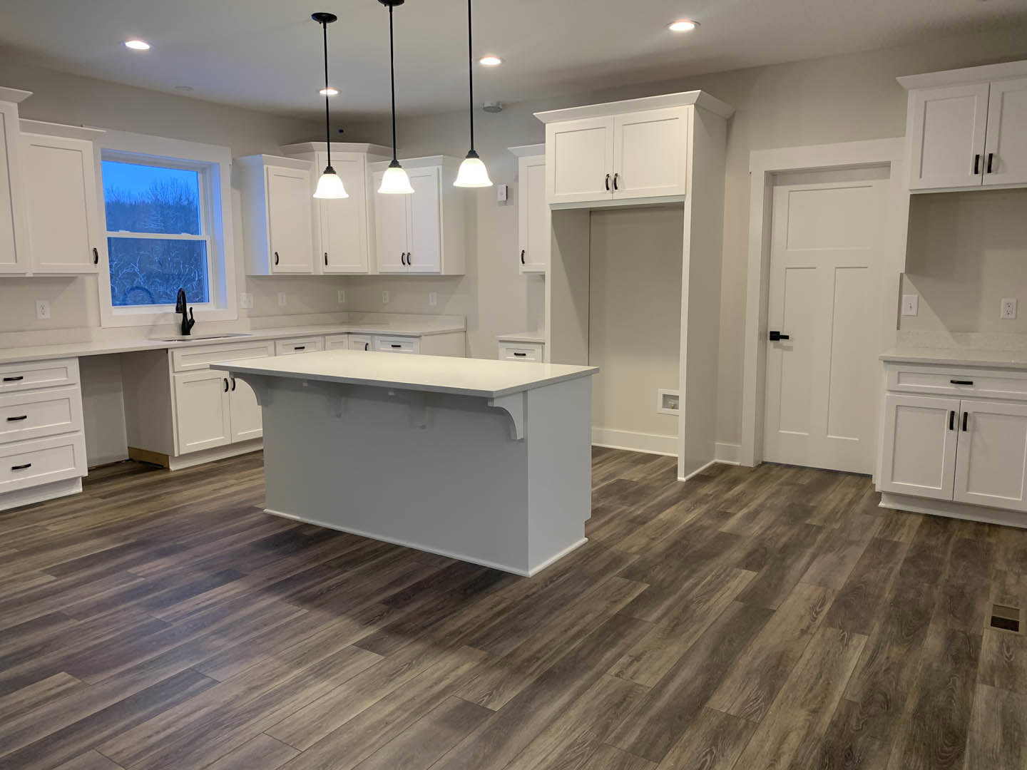 White kitchen cabinets with black handles, white countertop, wood flooring, large window overlooking trees, white walls, stainless steel sink, modern appliances