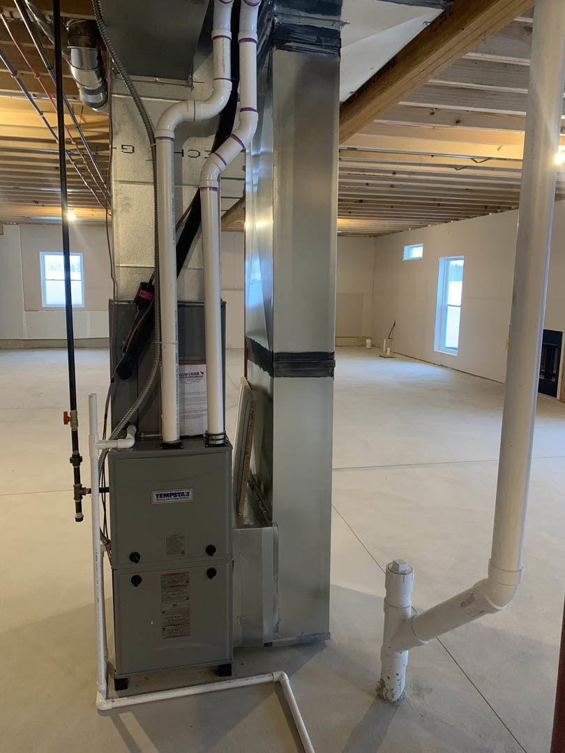 Spacious utility room featuring a large metal HVAC unit with exposed steel pipes, white painted walls, and a window allowing natural light onto the floor.