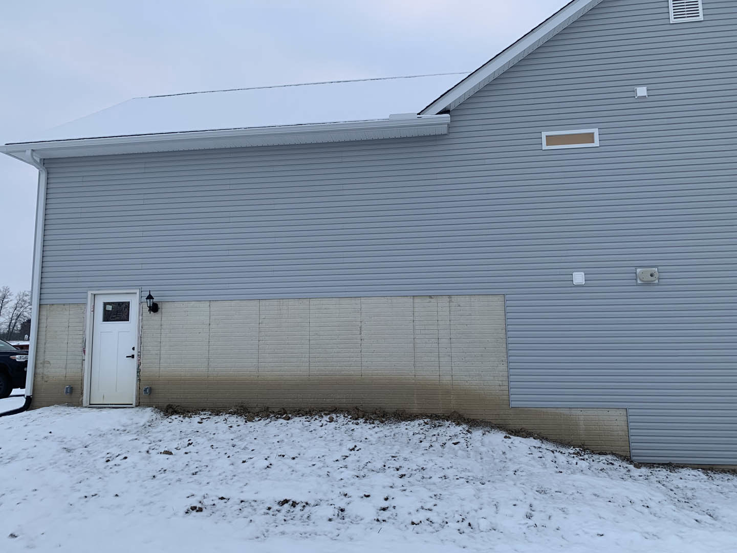 White brick house with blue roof, white door featuring black handle, snow covering ground, close-up window and tire visible in foreground