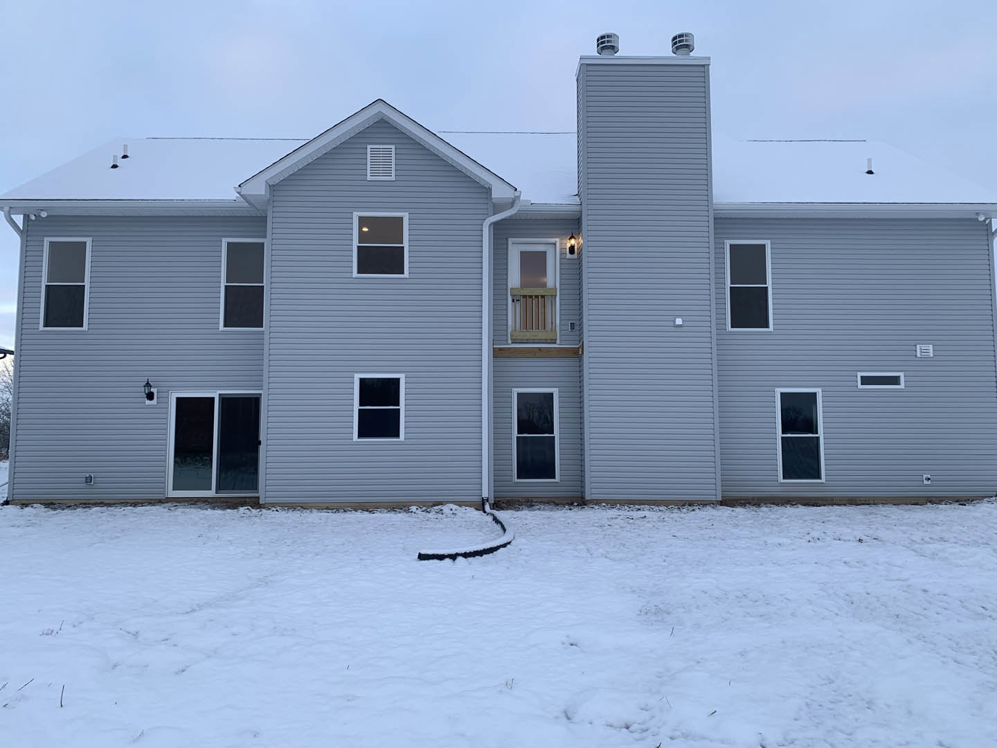 Grey siding house with white trim and chimney, snow covering ground, black drainage tube near foundation, Rockingham Meeting House visible in background.
