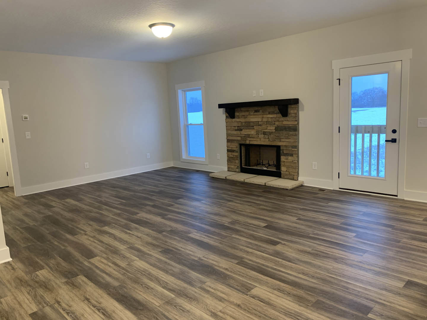 Living room with hardwood floors, central stone fireplace, black-framed accent chair, large window showing snowy landscape, modern ceiling light fixture