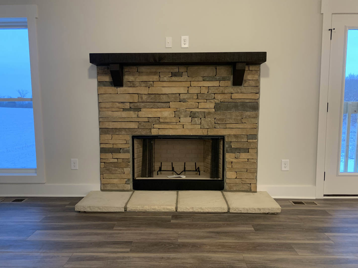 Black-framed fireplace with metal accents set in a white wall, wood flooring, large window overlooking snowy landscape