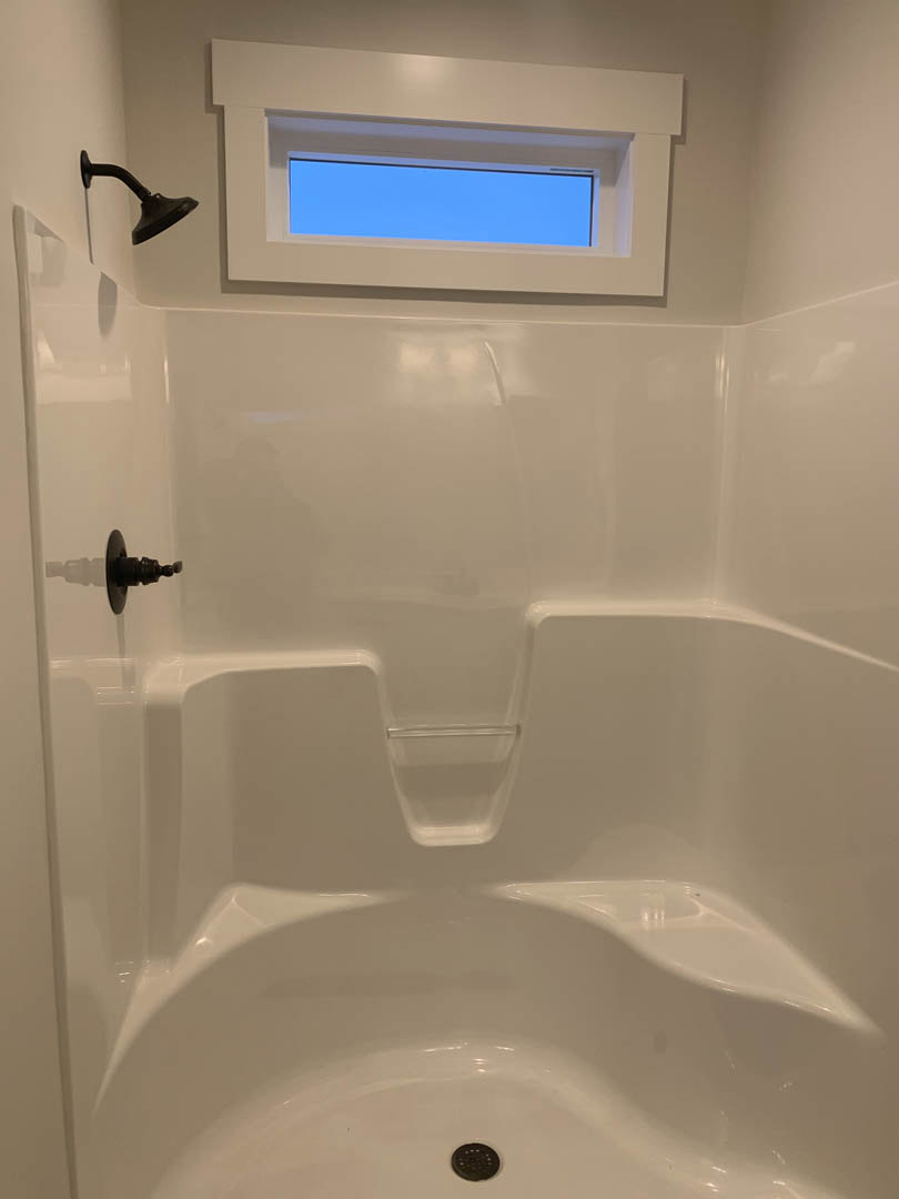 Shower with large window, white tile walls, black light fixture, white bathtub beneath window, blue sky visible outside