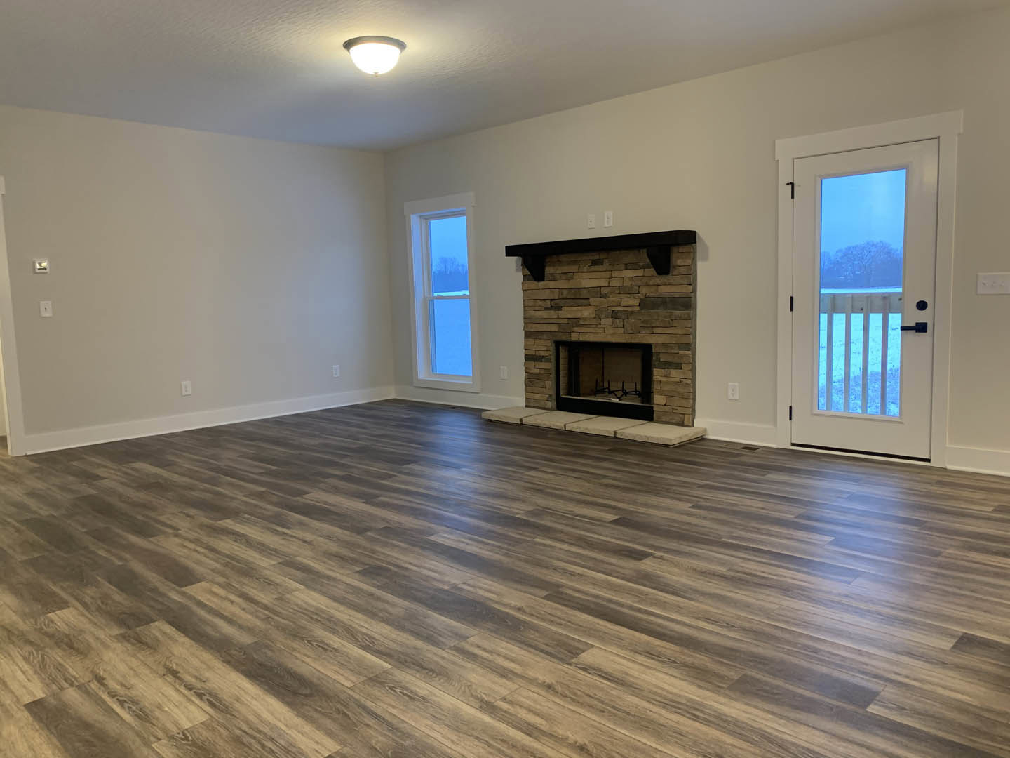 Living room with hardwood floors, white walls, black-framed fireplace with mantel, large window showing outdoor view, glass-paneled door, and ceiling light fixture.