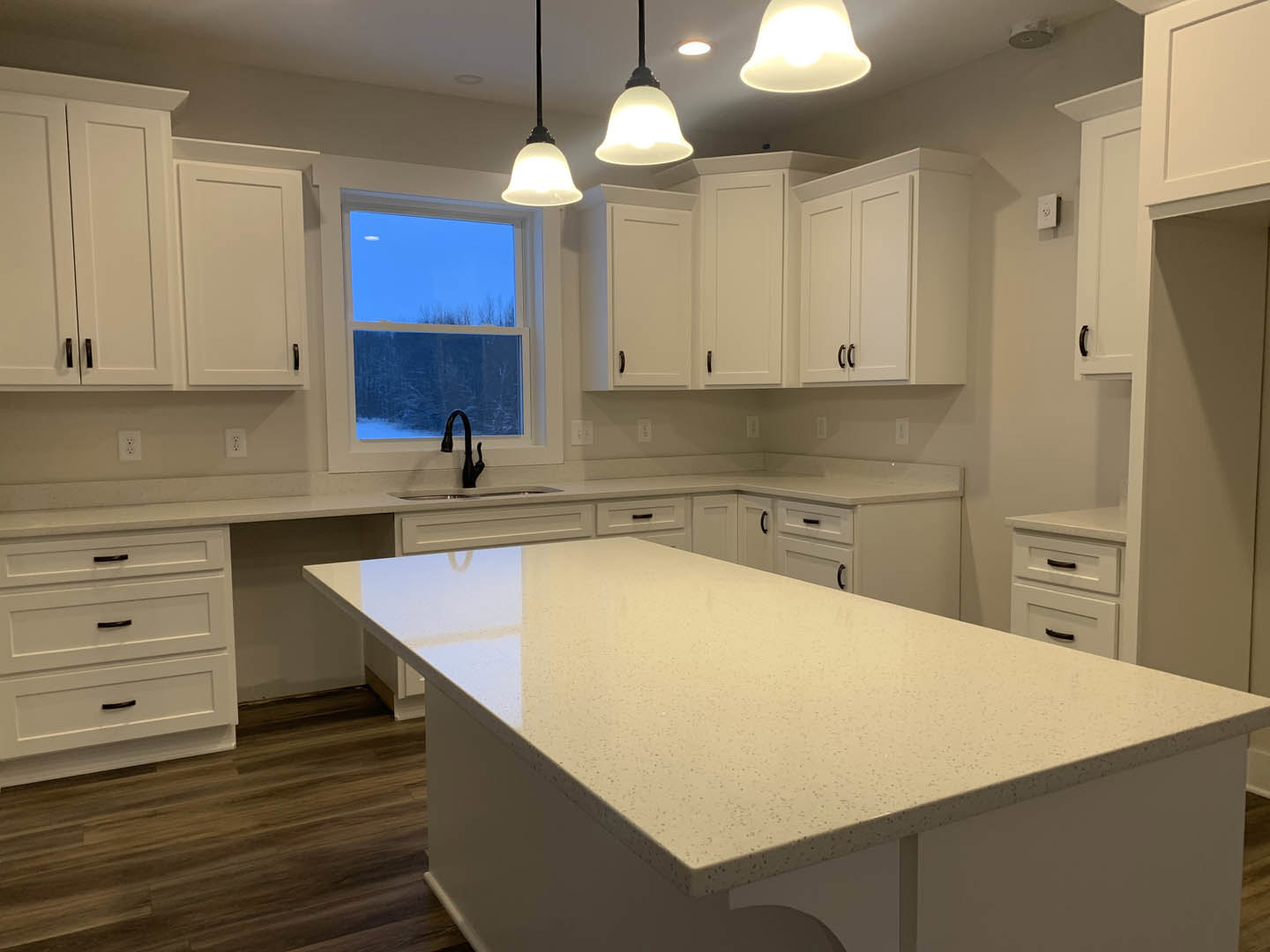 White kitchen with shaker cabinets, large central island featuring white quartz countertop, black curved faucet at undermount sink, wide window showing blue sky, white drawers and