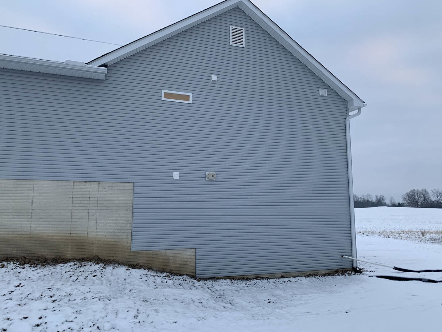 Modern house exterior with white siding, large garage door, and snow-covered ground; leafless trees in background, visible wall vent, overcast winter sky