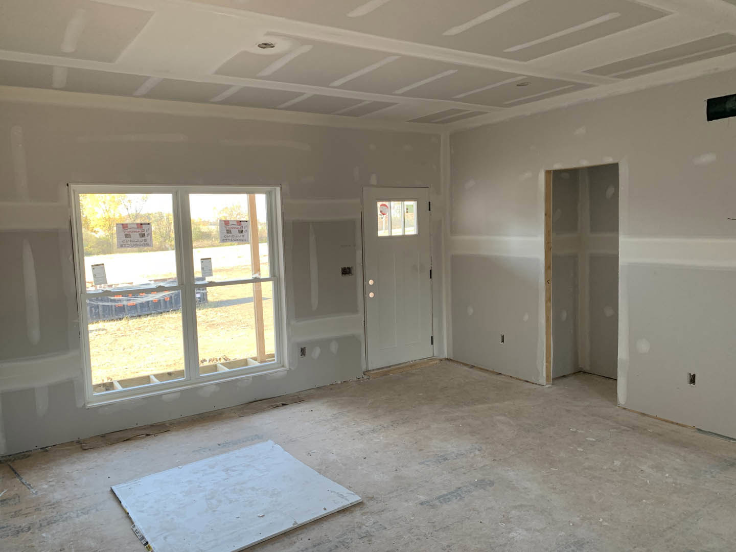 Bright room with light-colored plaster walls, wood flooring, white door, large windows displaying temporary signs, and minimal furnishings
