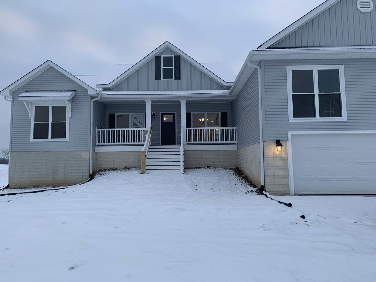 Two-story house with white siding, black front door illuminated by porch light, white-framed windows, attached garage with closed door, snow covering ground and steps leading to