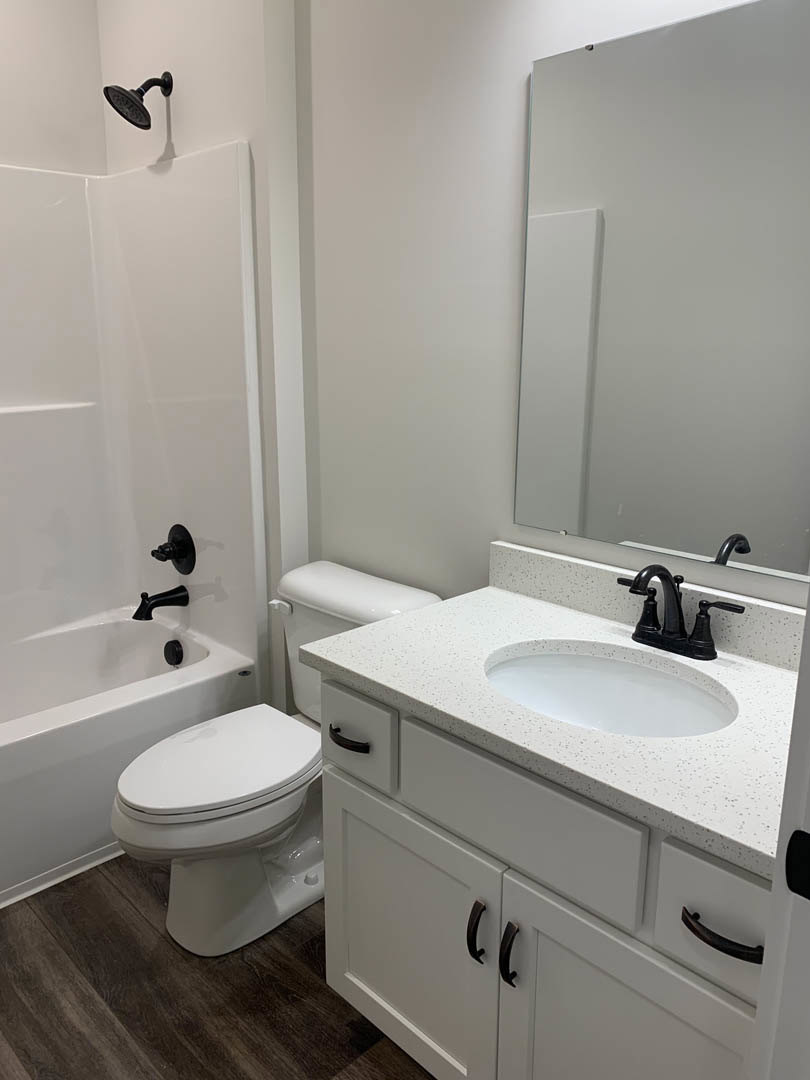 White bathroom with a freestanding bathtub, white toilet, white sink set in a stone countertop, matte black faucet, and light tile walls.