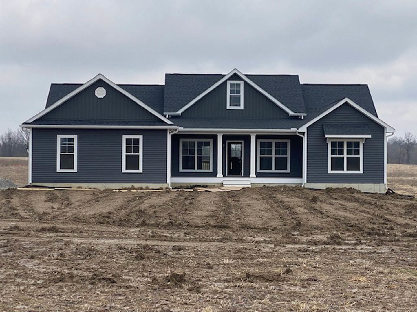 Two-story house under construction with grey roof, white-framed windows, light siding, exposed dirt yard, and person standing near window