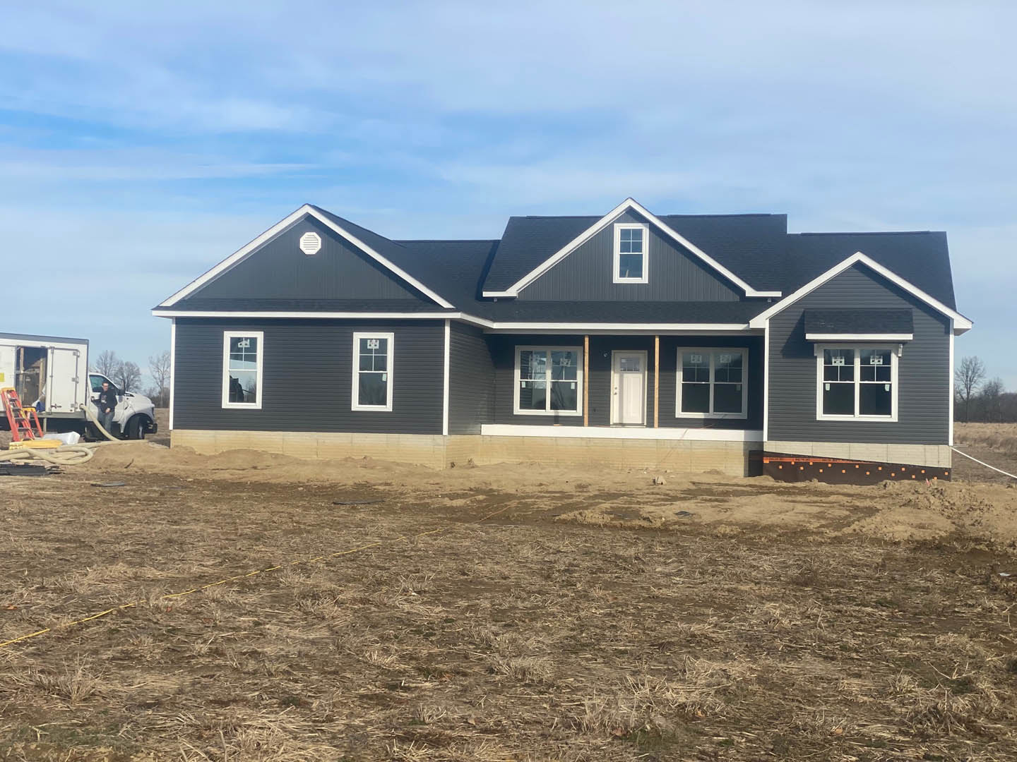 Partially built house with exposed framing, white door, white-framed windows, grassy and dirt yard, pickup truck parked in background, man standing nearby