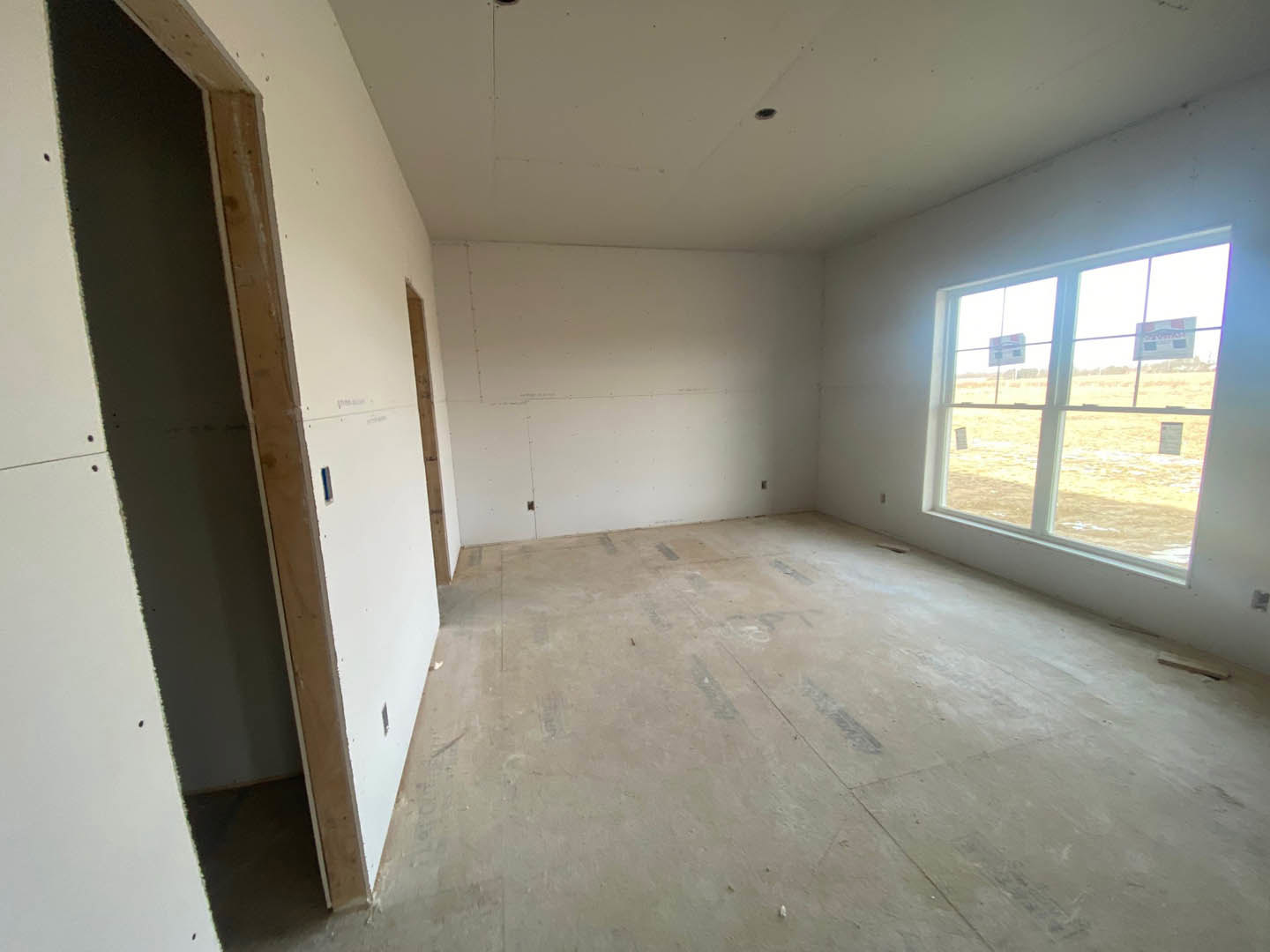 Room with unfinished plaster walls, concrete floor, large window featuring posted signs, and visible ceiling spot.