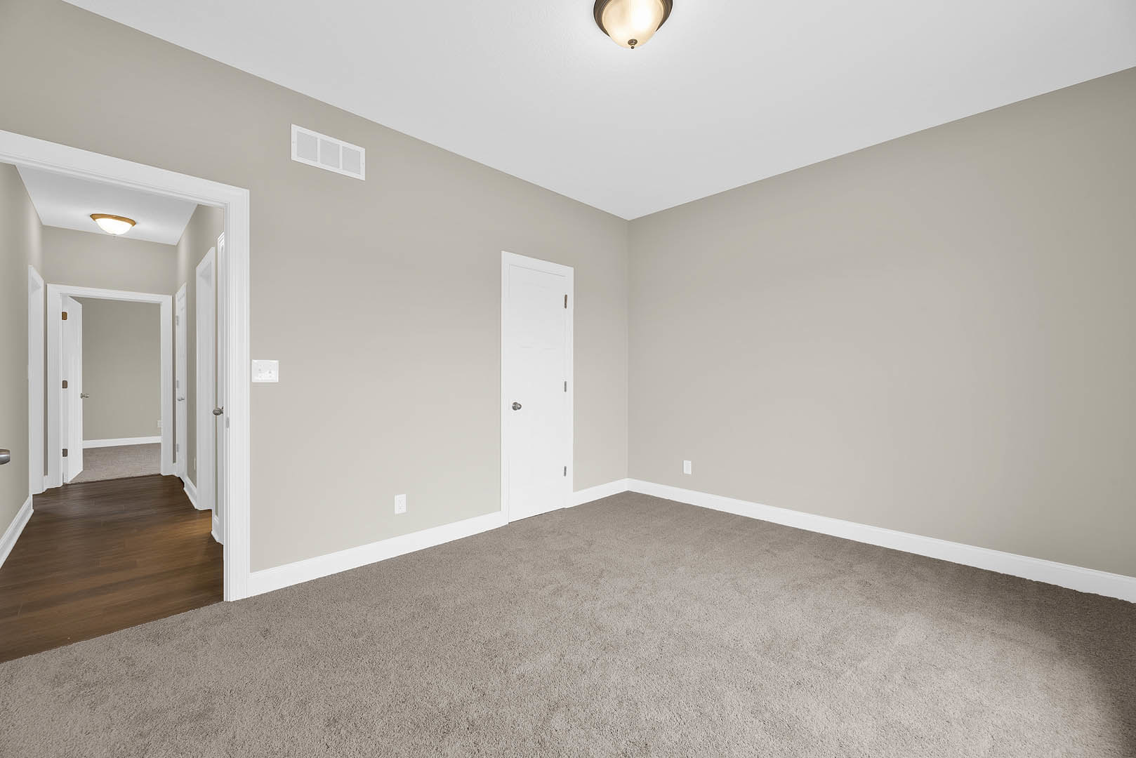 Carpeted room with white walls, white door featuring a silver knob, white ceiling vent, close-up of ceiling light, hardwood floor with white trim molding