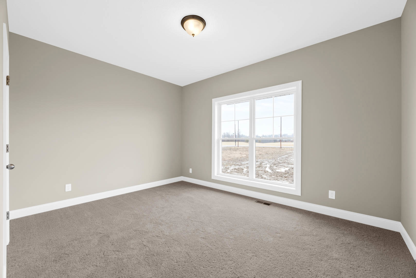 Neutral-toned carpeted room with white walls, large window overlooking muddy field, ceiling-mounted light fixture, simple molding along baseboards