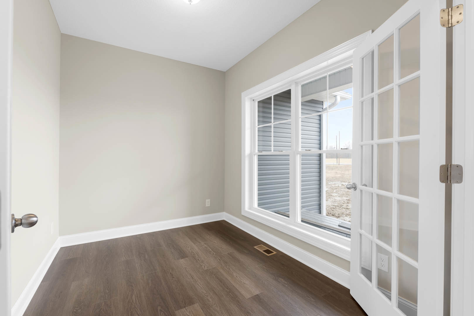 Hardwood floor with white baseboard trim, white walls, large window with divided glass panes, white door featuring glass panels and metal knob, close-up of door hinge