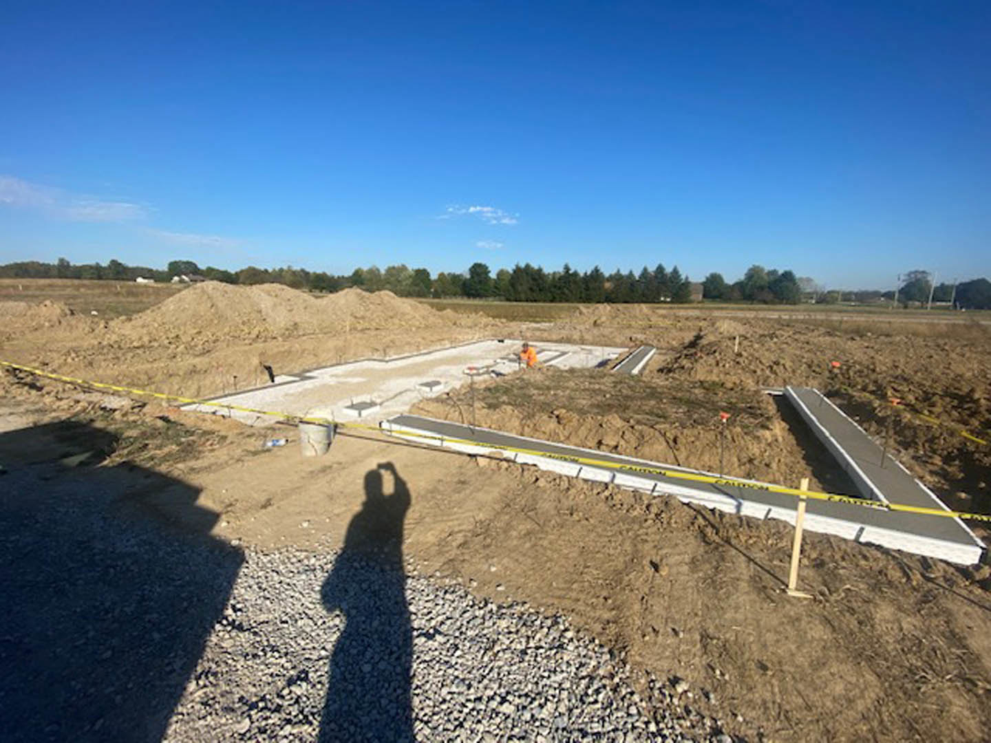 Gravel construction site with a large dirt pile, scattered soil, and a man photographing under a blue sky with clouds; tree and plant visible along road.